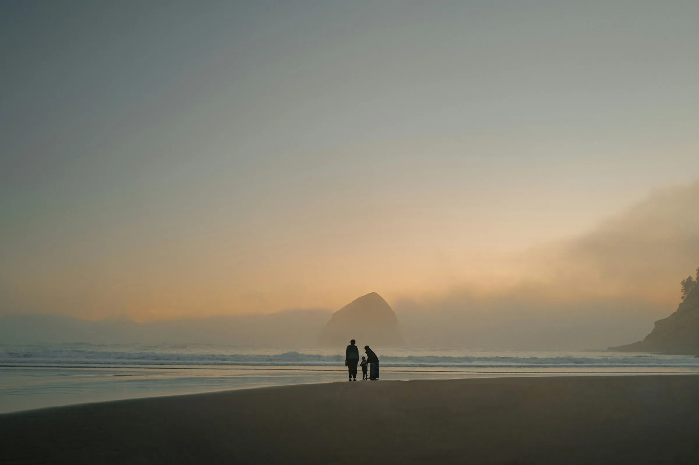 Two small figures on a wide beach in sea fog at sunset, a sea stack behind them.