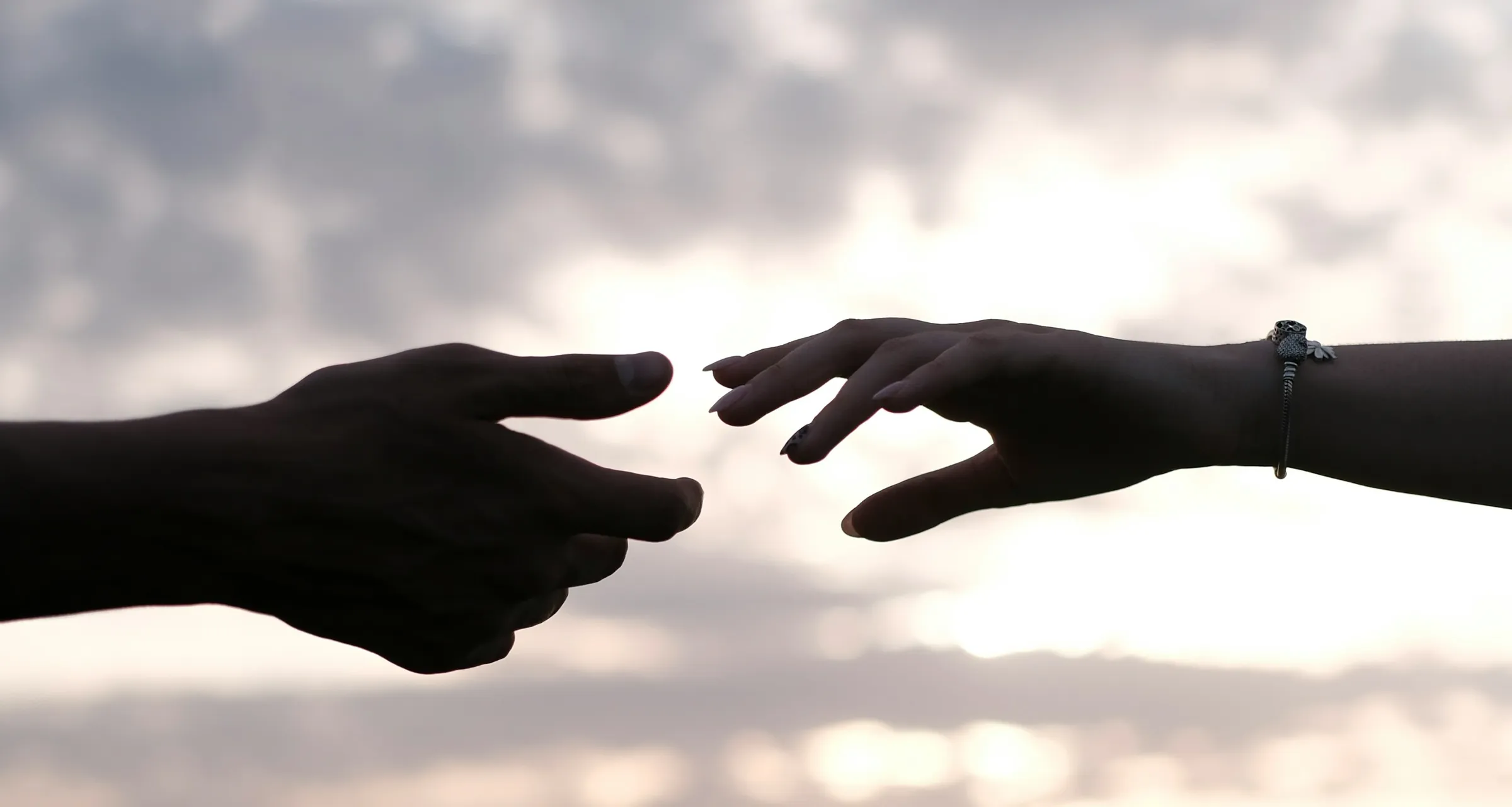 Two hands reaching toward each other in silhouette against an evening sky, almost touching.